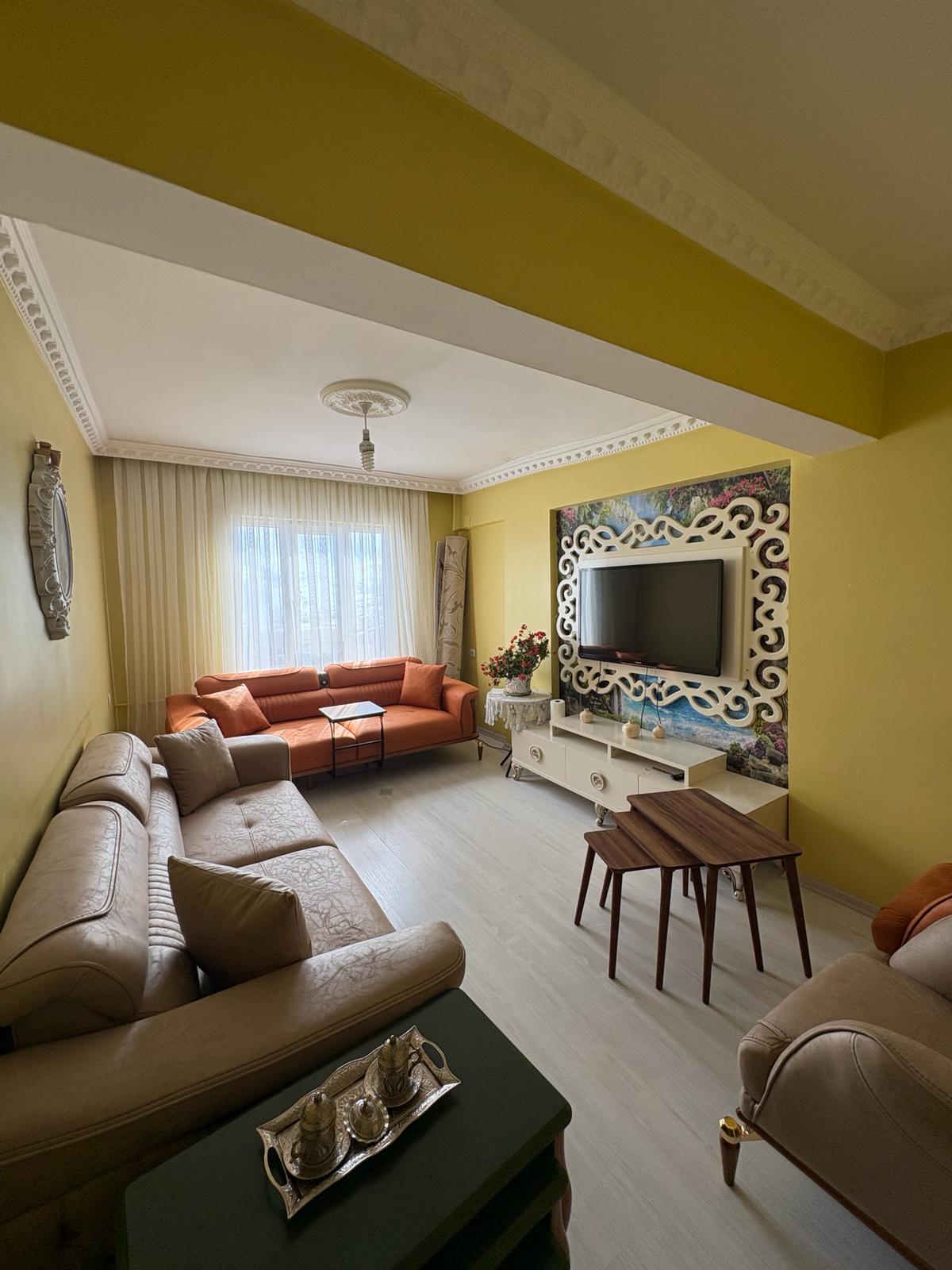 Beige corner and orange three-seater sofas from the Gülistan R. 2026 collection in a modern living room with yellow walls, featuring Inegol furniture quality.