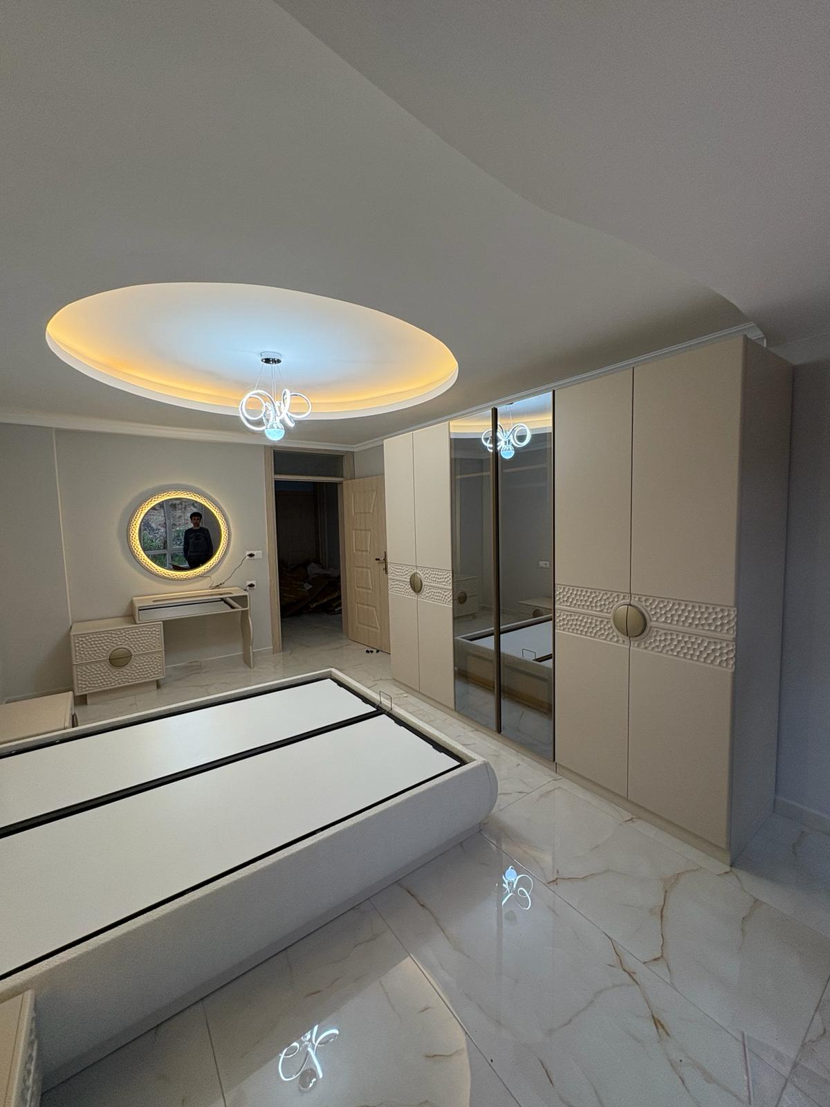 Modern Mehmet Emin G. 2026 beige bedroom set featuring a mirrored wardrobe, elegant dressing table, and stylish lighting. A prime example of Inegol furniture models.