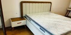 Modern bir yatak odasında yer alan, bej döşemeli yatak başlığı, beyaz bazalı yatak ve ahşap-beyaz komodinler. Yeni ve şık bir İnegöl mobilyası örneği.