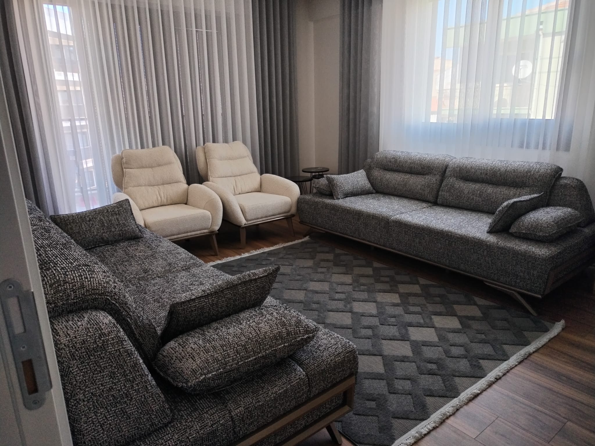 A modern and elegant living room set featuring a grey L-shaped sofa, a three-seater sofa, and two cream armchairs. This Affordable İnegöl furniture set is completed with a patterned rug and bright curtains.