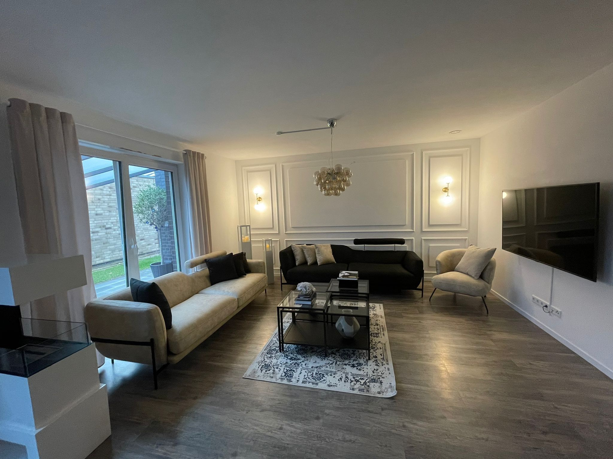 A spacious and bright living room featuring stylish modern Inegol furniture in beige and black, complemented by large windows and contemporary lighting.