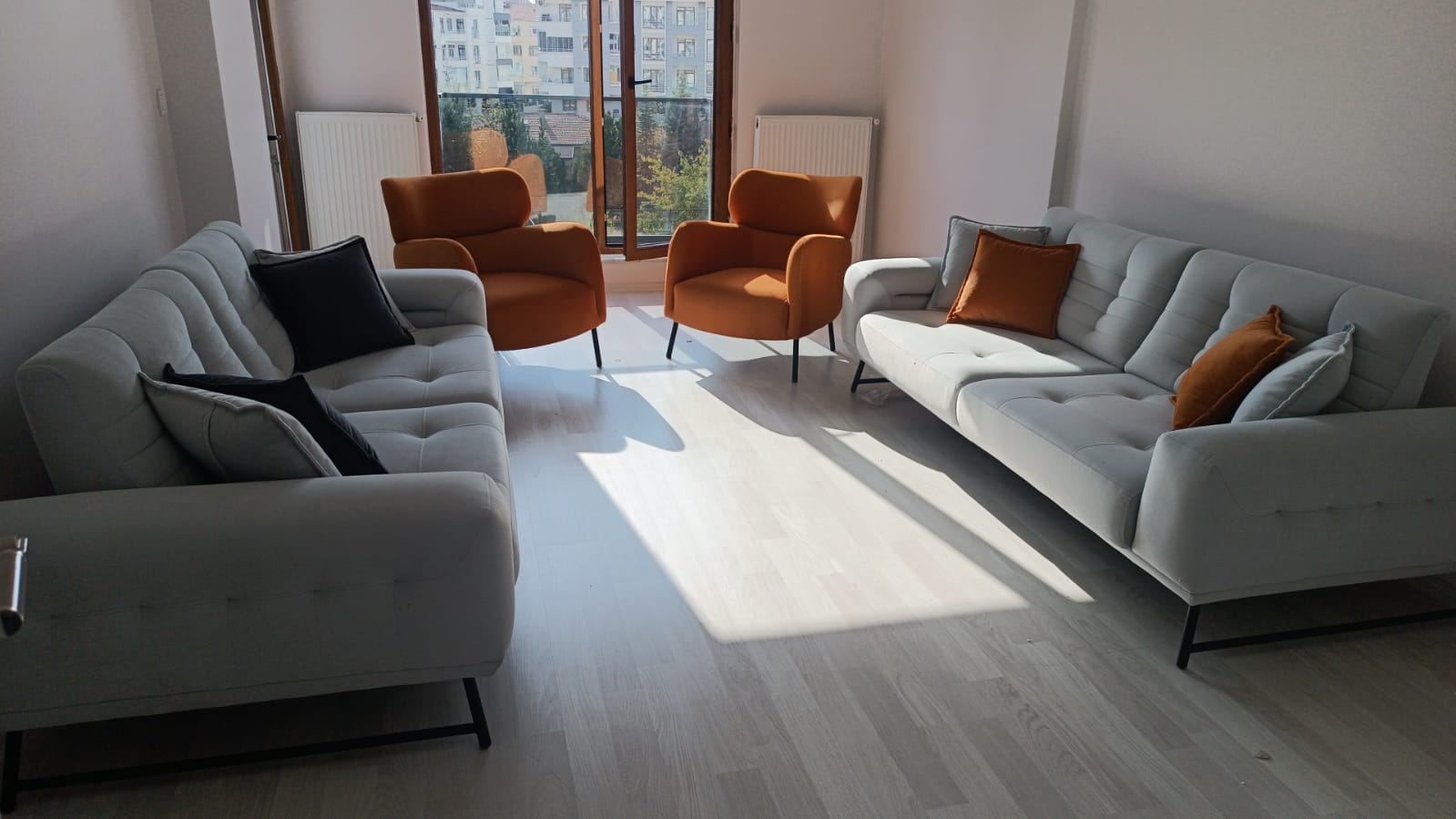 A stylish modern living room set featuring two light gray sofas, two vibrant orange armchairs, and matching throw pillows under natural light. Explore Inegol Furniture Prices.