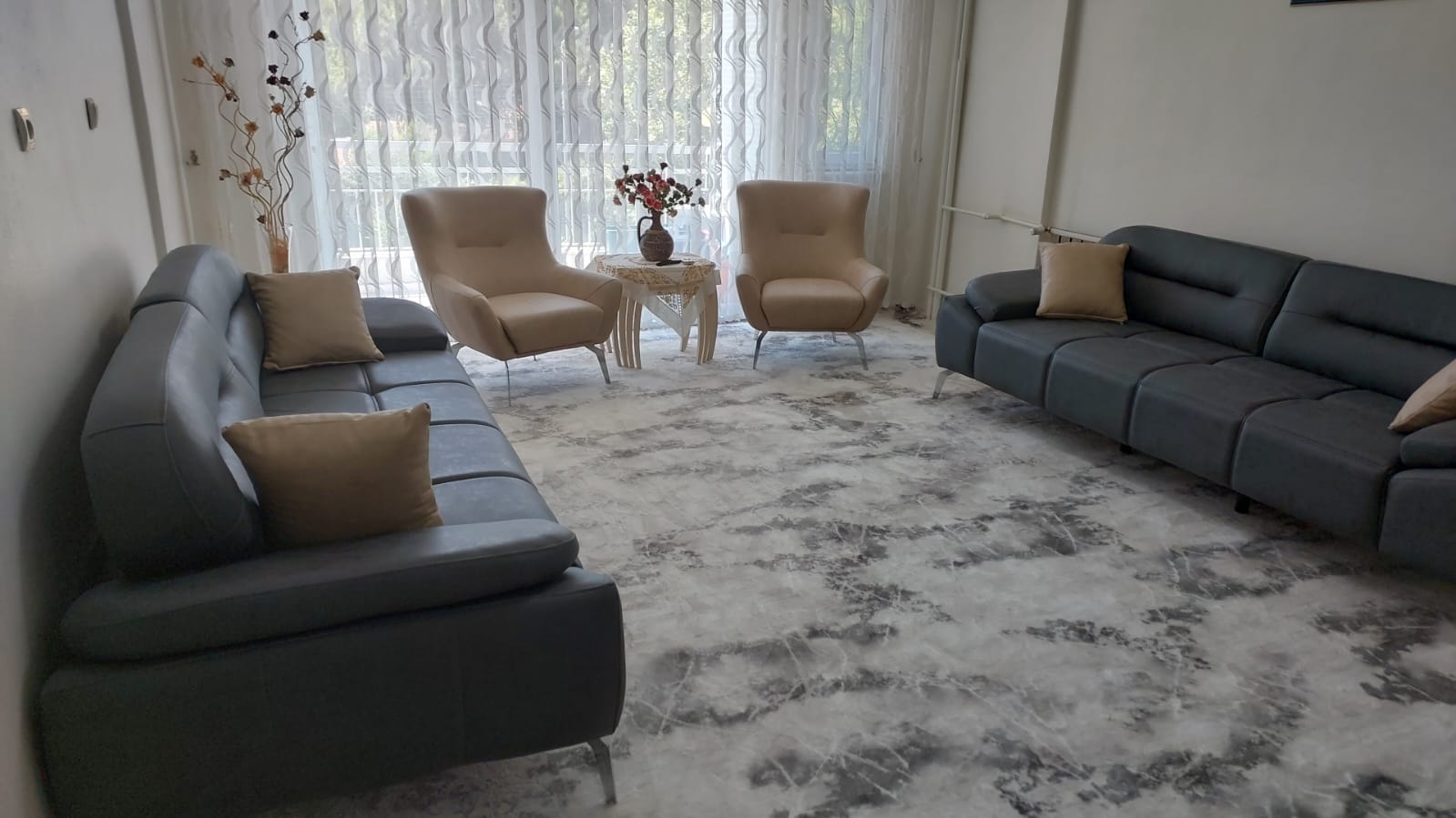 Spacious living room featuring a dark grey modern Inegol furniture sofa set, beige armchairs, marble pattern rug, and elegant decor.