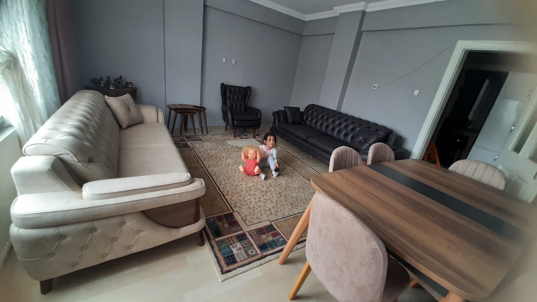 Child playing with a doll in a spacious living room featuring beige and black sofas, a wooden dining table, and quality inegol furniture.