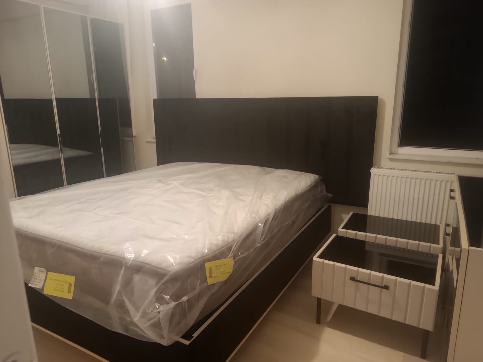 Stylish bedroom set with dark grey upholstered bed frame, new mattress in wrapping, and two elegant white mirrored nightstands. Affordable Inegol furniture.