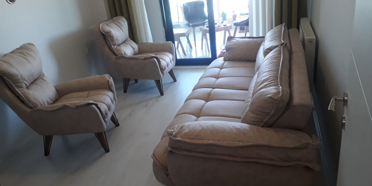 Balkon manzaralı bir odada, bej renkli modern inegol mobilya tarzında üçlü kanepe ve iki tekli berjerden oluşan şık oturma grubu.